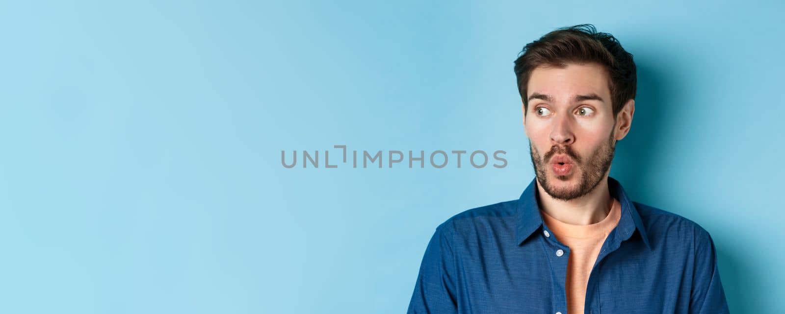 Close up of impressed man say wow and checking out banner, looking left at empty space amazed, standing on blue background by Benzoix