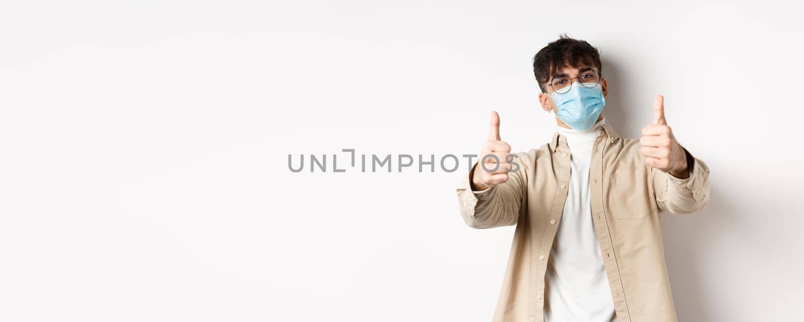 Coronavirus, health and real people concept. Smiling guy in medical mask showing thumbs up, wearing glasses, standing on white background.