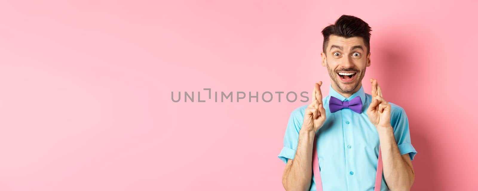 Happy guy making wish, holding fingers crossed for good luck and smiling at camera, waiting for good news, standing over pink background by Benzoix