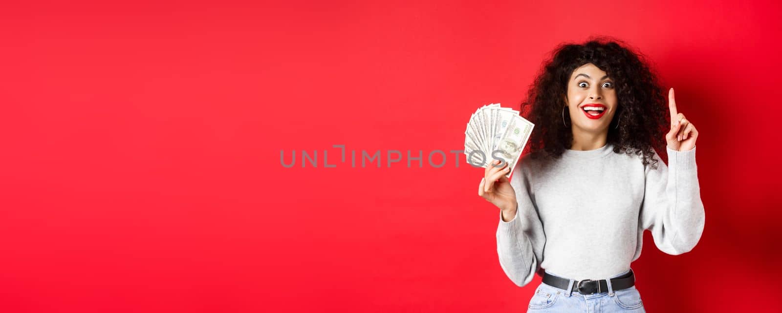 Young excited woman having an idea how make money, showing dollar bills and raising finger in eureka sign, standing on red background by Benzoix