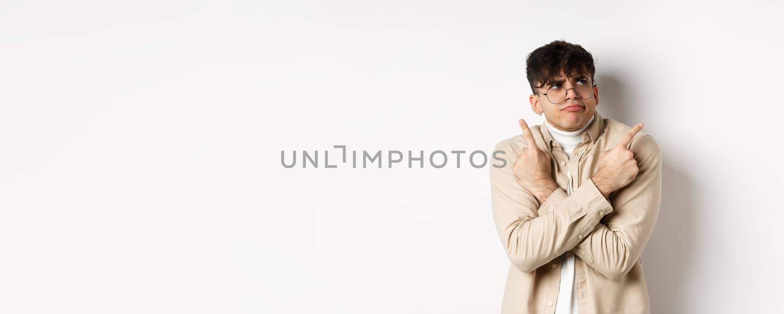 Indecisive funny young man having troubles with making choice, pointing fingers sideways and frowning perplexed, deciding between two variants, standing on white background.