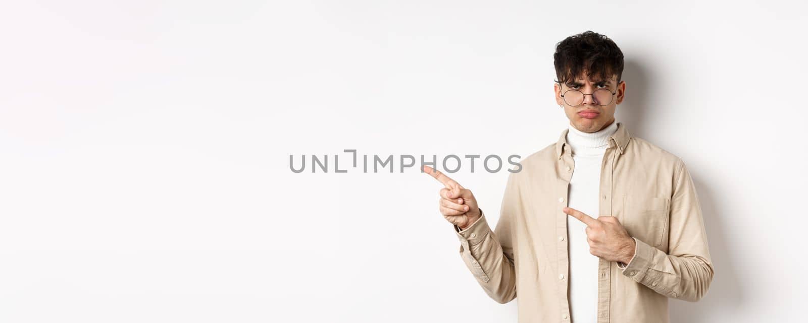 Sad and grumpy boy pointing fingers left, sulking with childish expression, complaining on something bad, standing on white background by Benzoix