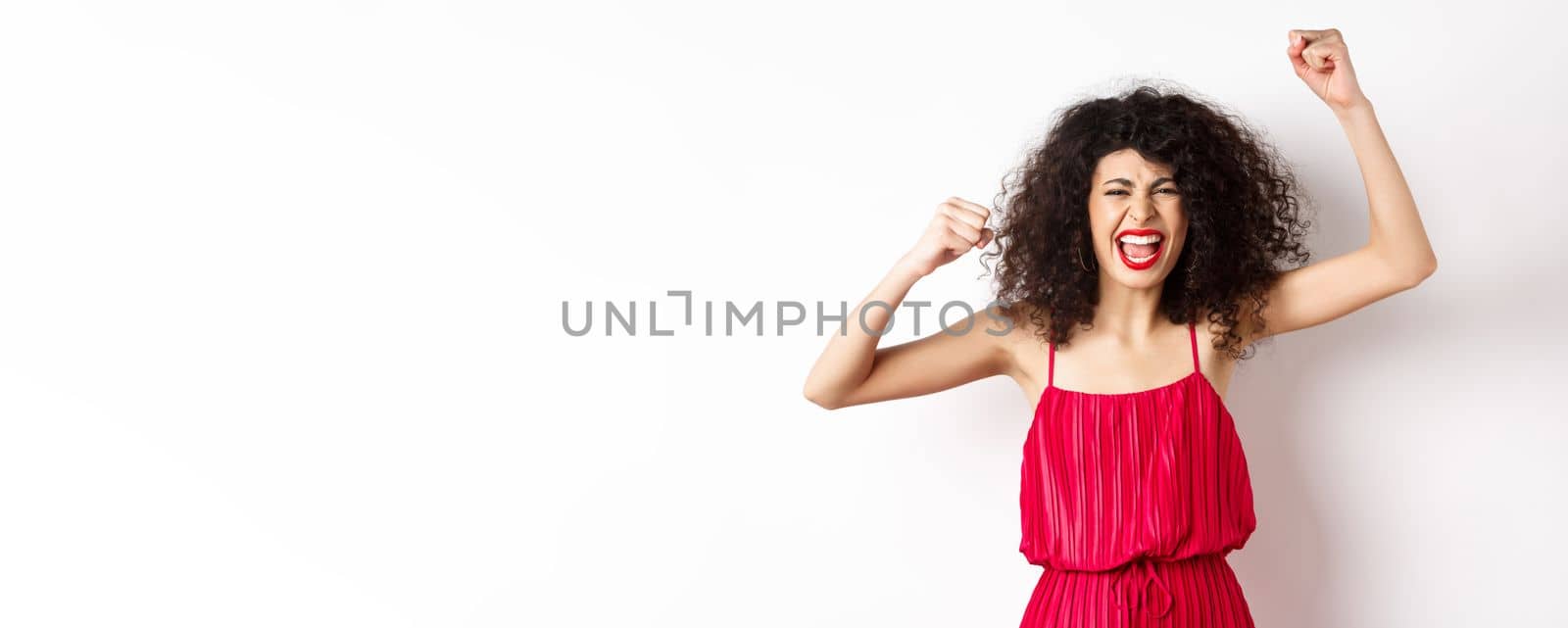Cheerful emotive woman with curly hair, red dress, raising hands up and chanting, rooting for team, shouting wanting to win, standing on white background by Benzoix