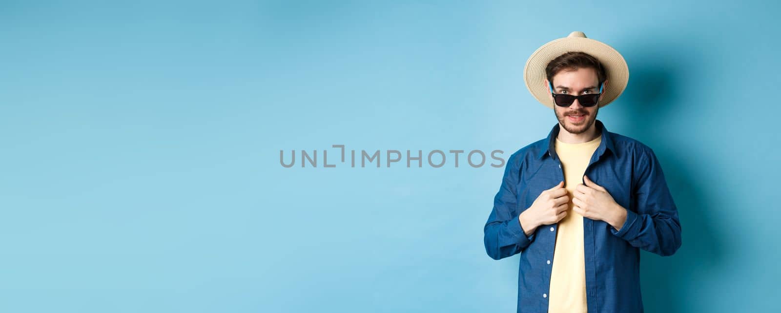 Handsome young man going on vacation, adjusting shirt, wearing sunglasses and straw hat. Concept of summer holiday.