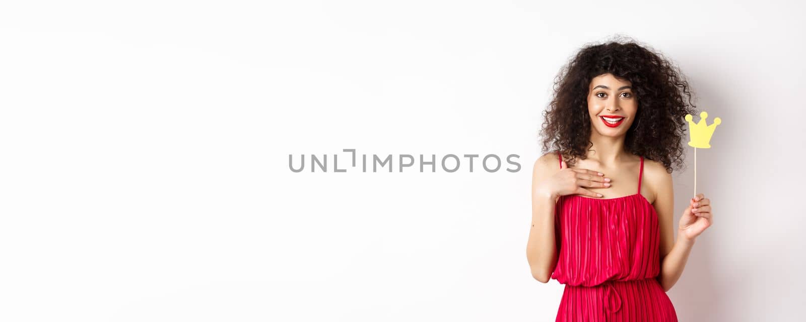 Confident pretty lady in red dress, holding queen crown on stick and looking excited, standing on white background.