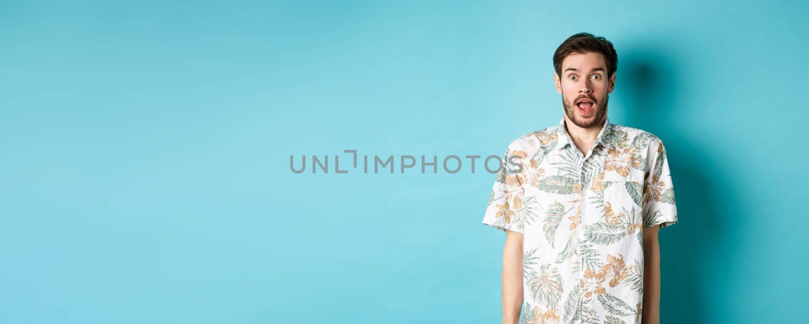 Summer holiday. Surprised guy in hawaiian shirt gasping amazed, checking out vacation offer, standing on blue background.