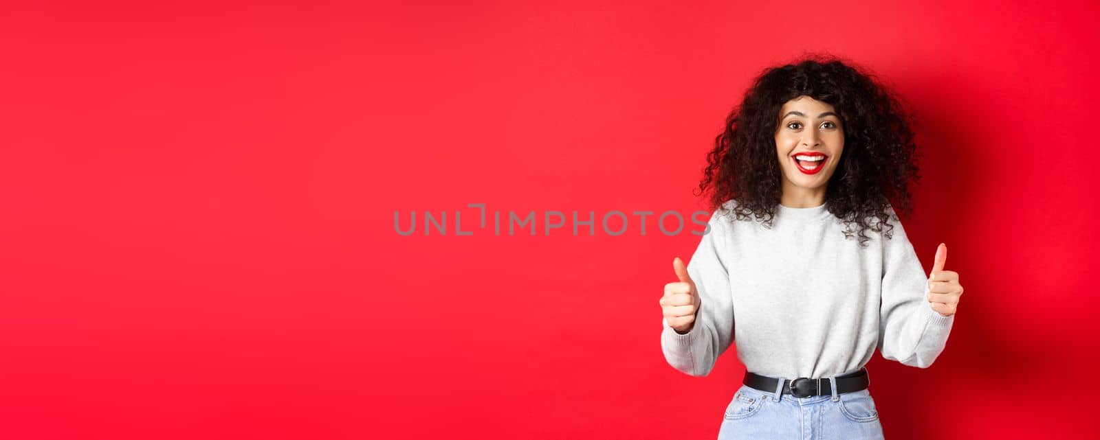 Enthusiastic girl with curly hair and red lips, showing thumbs up and saying yes, agree with you, compliment good work, like something cool, standing on studio background by Benzoix