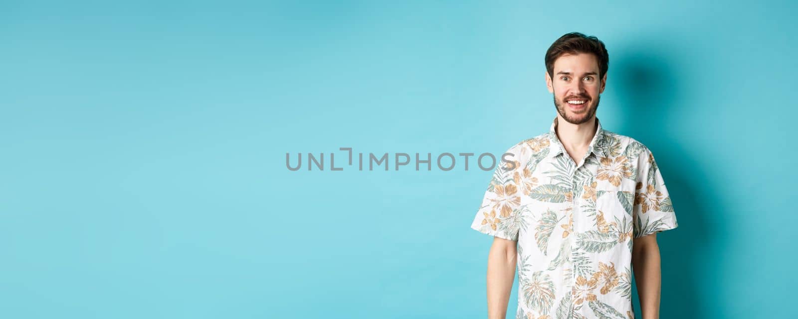 Cheerful caucasian guy going on vacation, wearing summer shirt and smiling, standing on blue background.