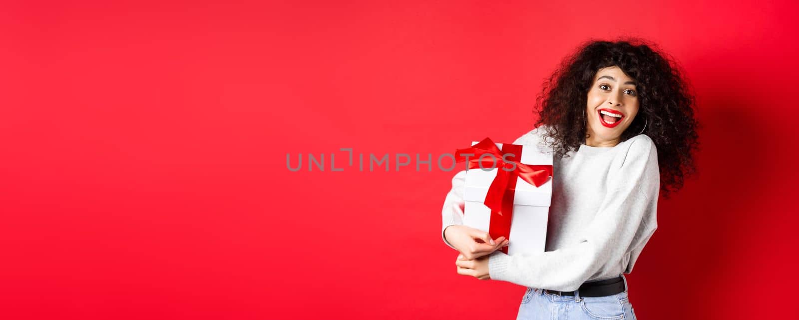 Valentines day. Happy girlfriend holding gift box from secret admirer, looking romantic at lover, receive present, standing on red background.