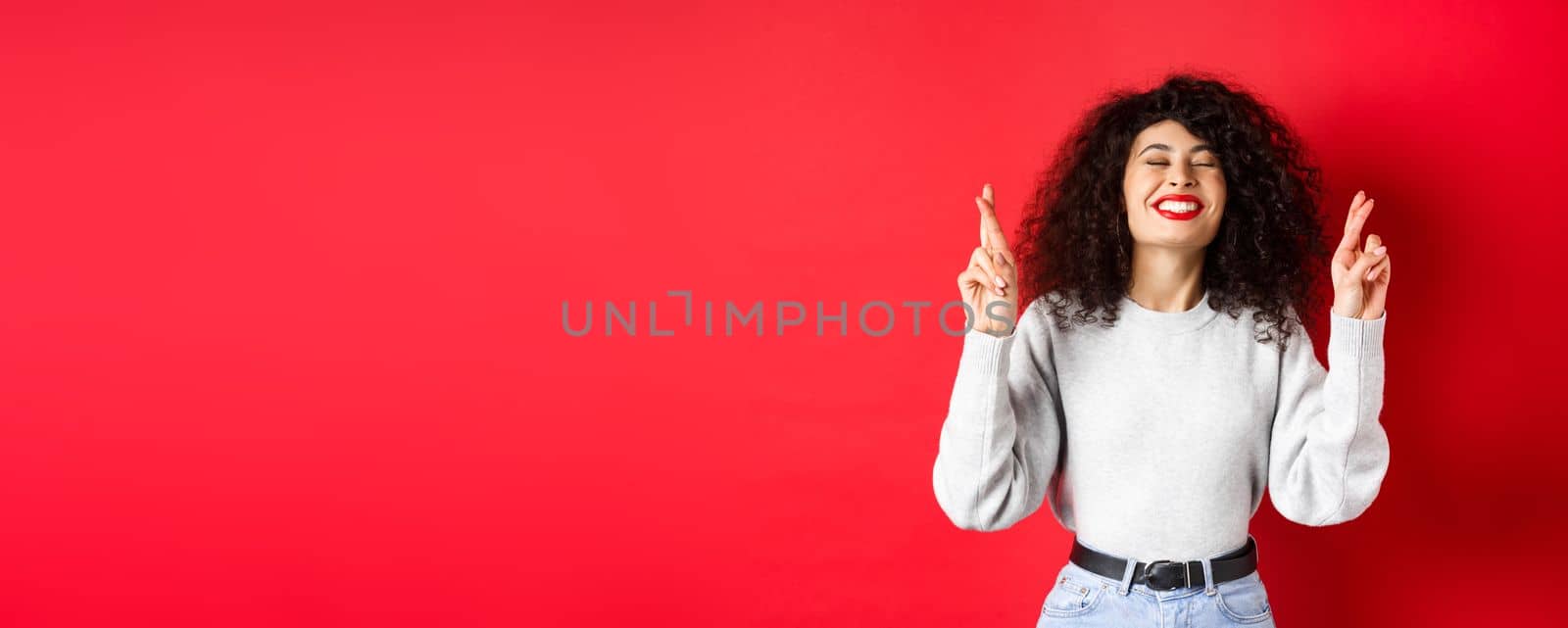 Hopeful and positive woman with red lips and curly hair, cross fingers for good luck and making wish, praying for dream come true, smiling excited, red background by Benzoix