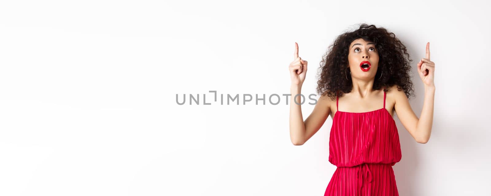 Amazed girl with evening makeup and curly hair, looking and pointing up excited, showing advertisement, standing in red dress on white background.