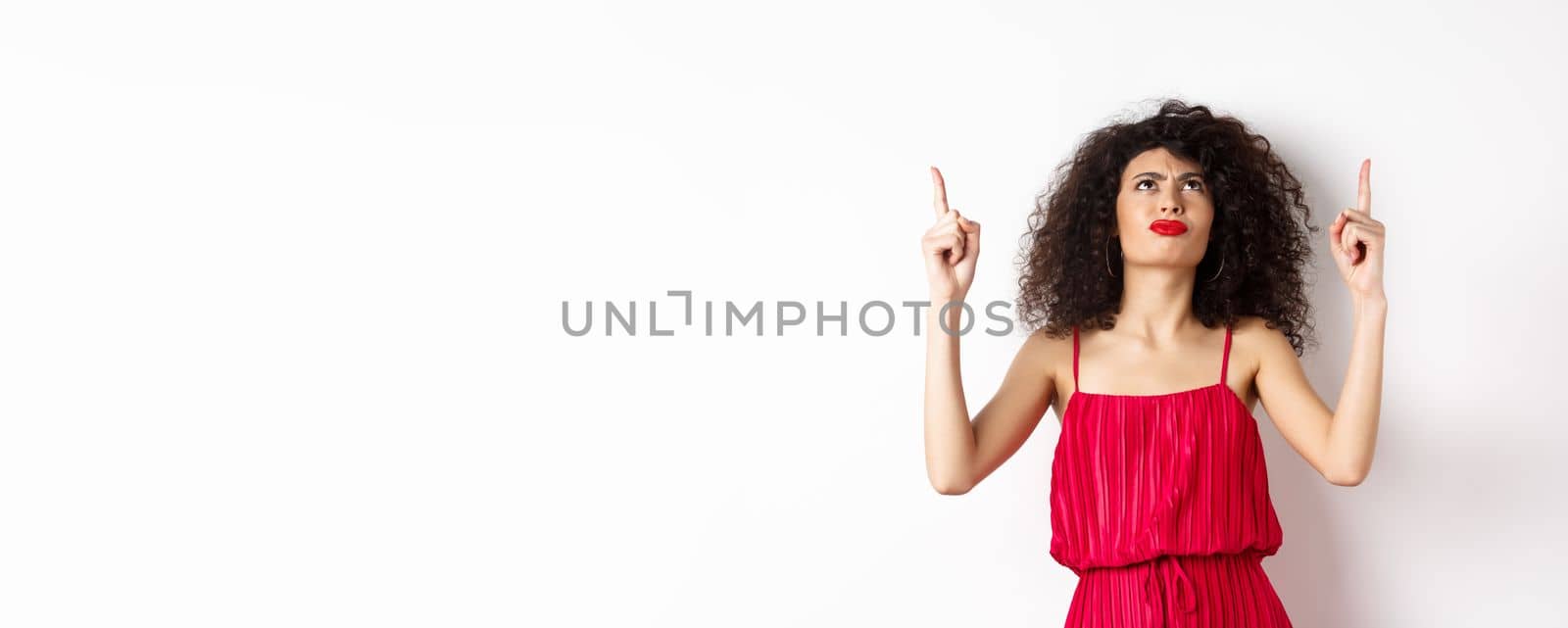 Angry and grumpy woman with curly hair, wearing red dress, frowning and looking up disappointed, standing over white background.