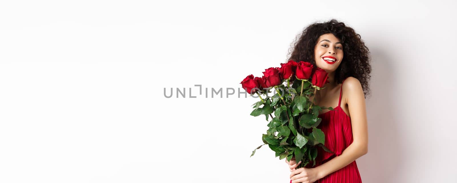 Romantic lady with curly hair and fashionable dress, holding bouquet of red roses and smiling, standing happy on white background.