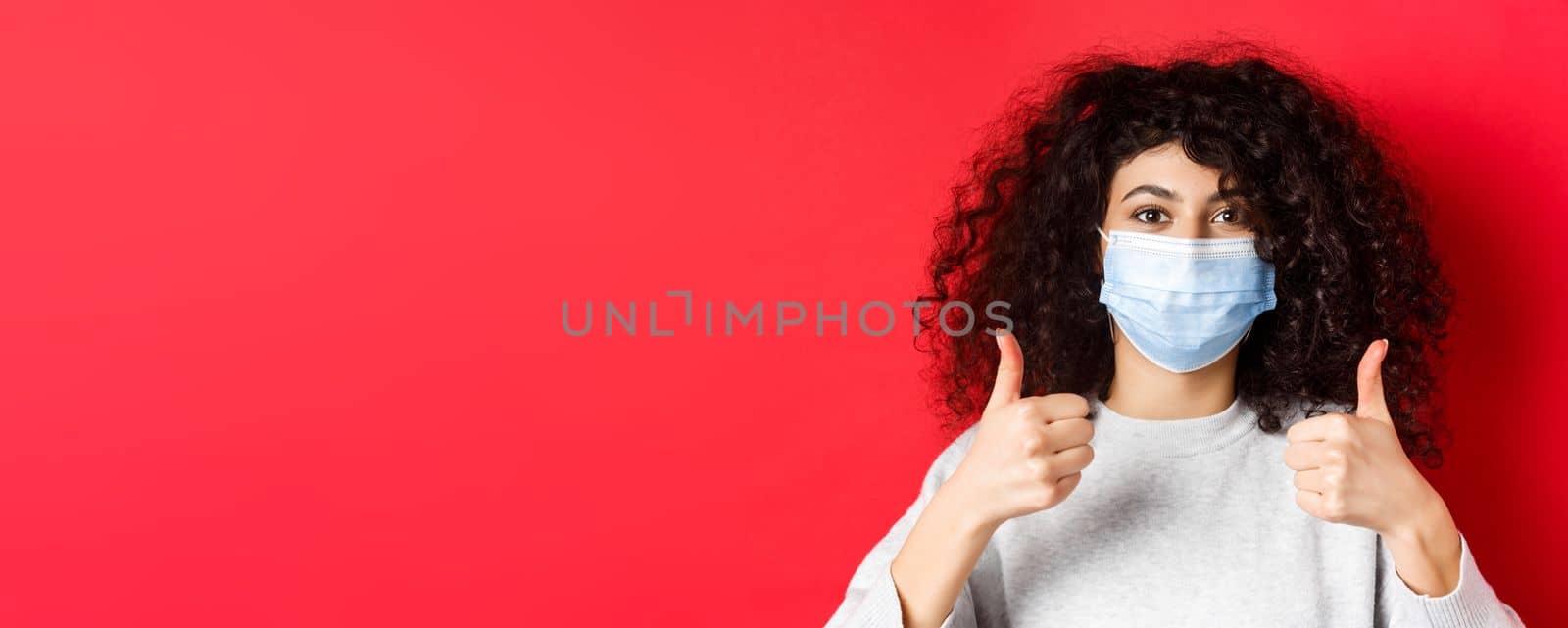 Covid-19 and pandemic concept. Close-up of young curly woman in face mask showing thumbs up, like and praise good thing, standing on red background.
