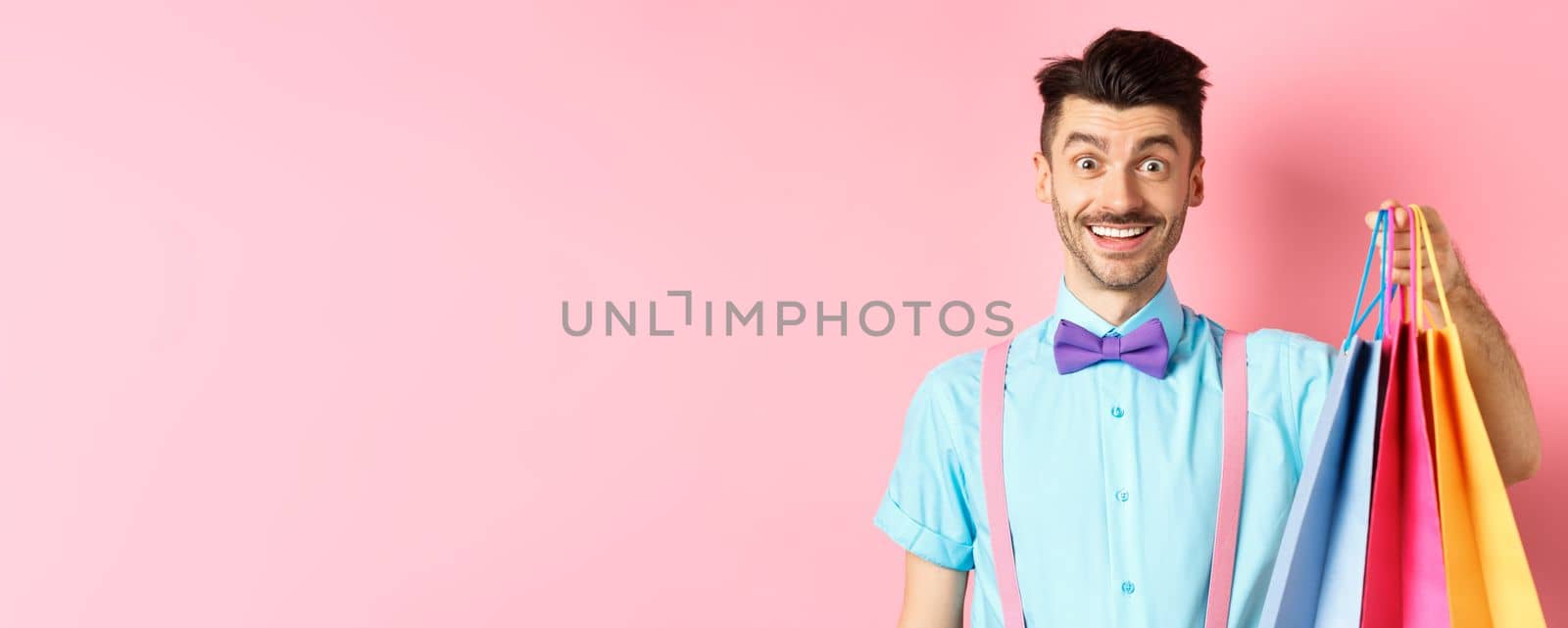 Image of happy guy on shopping, holding paper bags and smiling excited, shopper buying with discounts, standing on pink background.