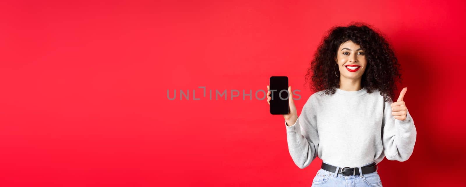 Portrait of attractive smiling woman with curly hair, showing empty mobile phone screen and thumb-up, recommending online promo, standing on red background.