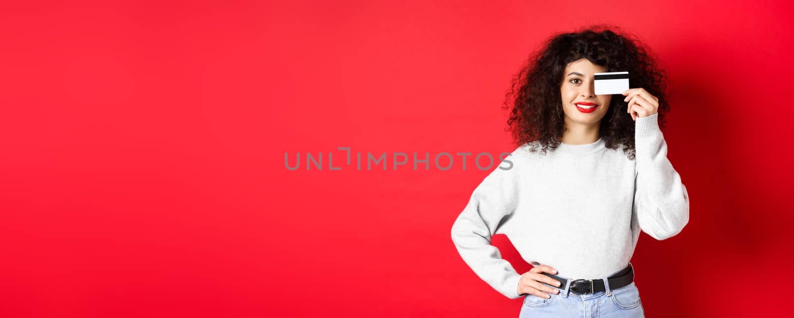 Beautiful female model with curly hair, showing plastic credit card and smiling, standing on red background. Makeup and shopping concept.