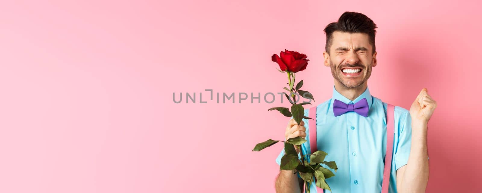 Romance and Valentines day concept. Hopeful man feeling excited before date, waiting for lover with red rose and praying, standing over pink background.