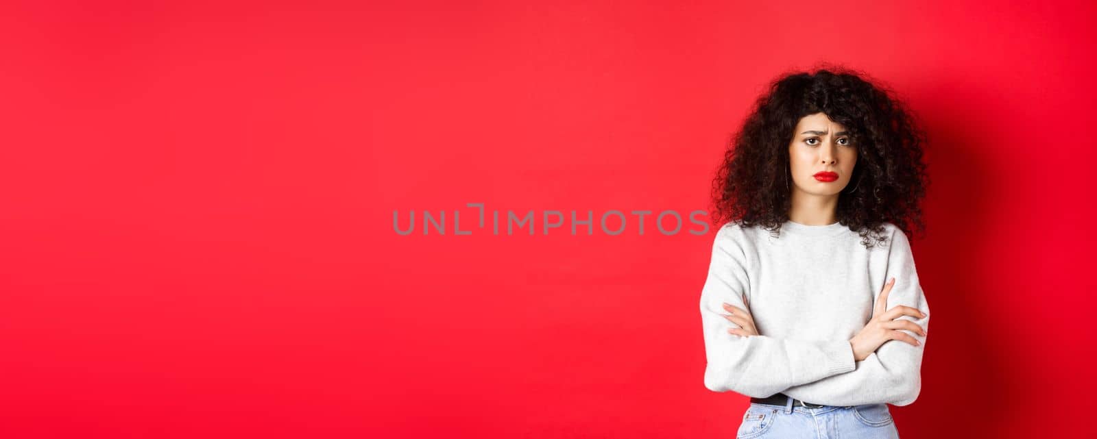 Sad and worried caucasian woman frowning, cross arms on chest and looking concerned, feeling bad, standing on red background.