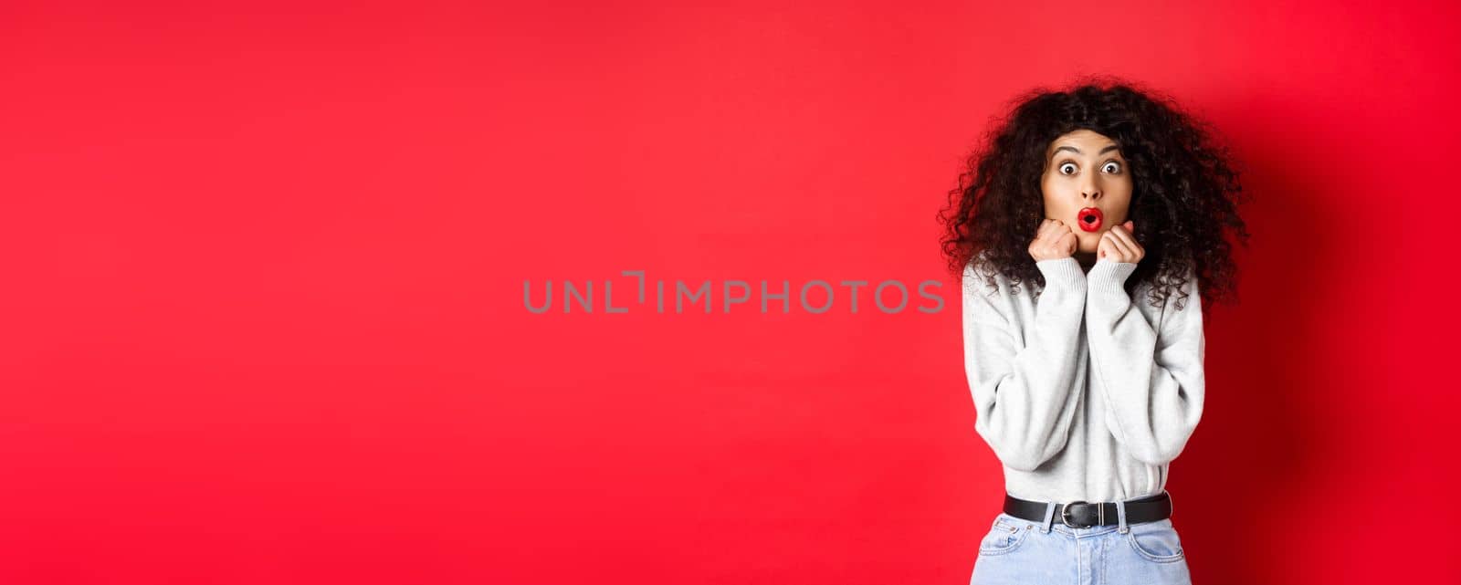 Surprised young woman looking with disbelief and amazement at camera, saying wow and standing in awe on red background.