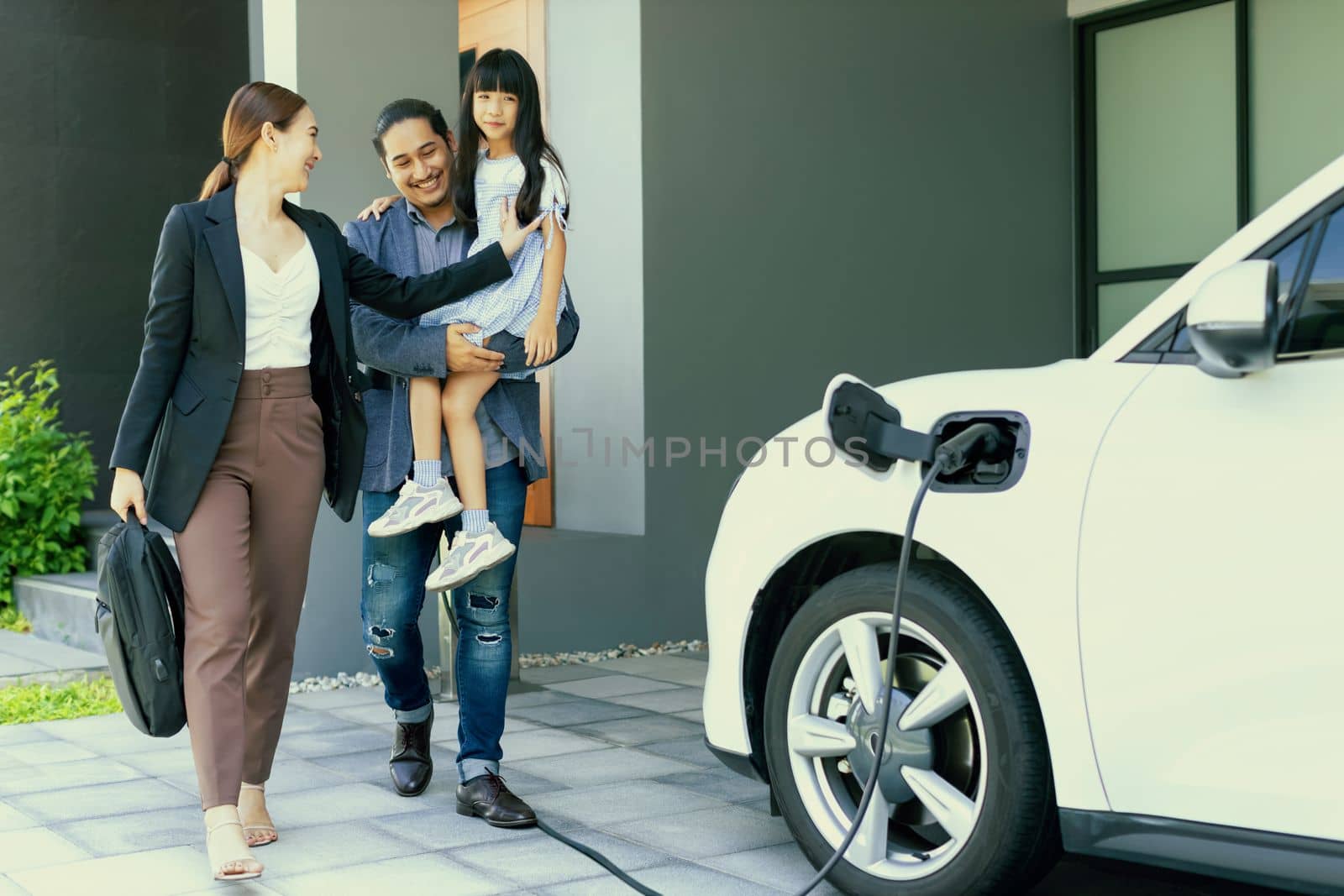 Progressive young parents and daughter with electric vehicle and home charging station. Green and clean energy from electric vehicles for healthy environment. Eco power from renewable source at home.