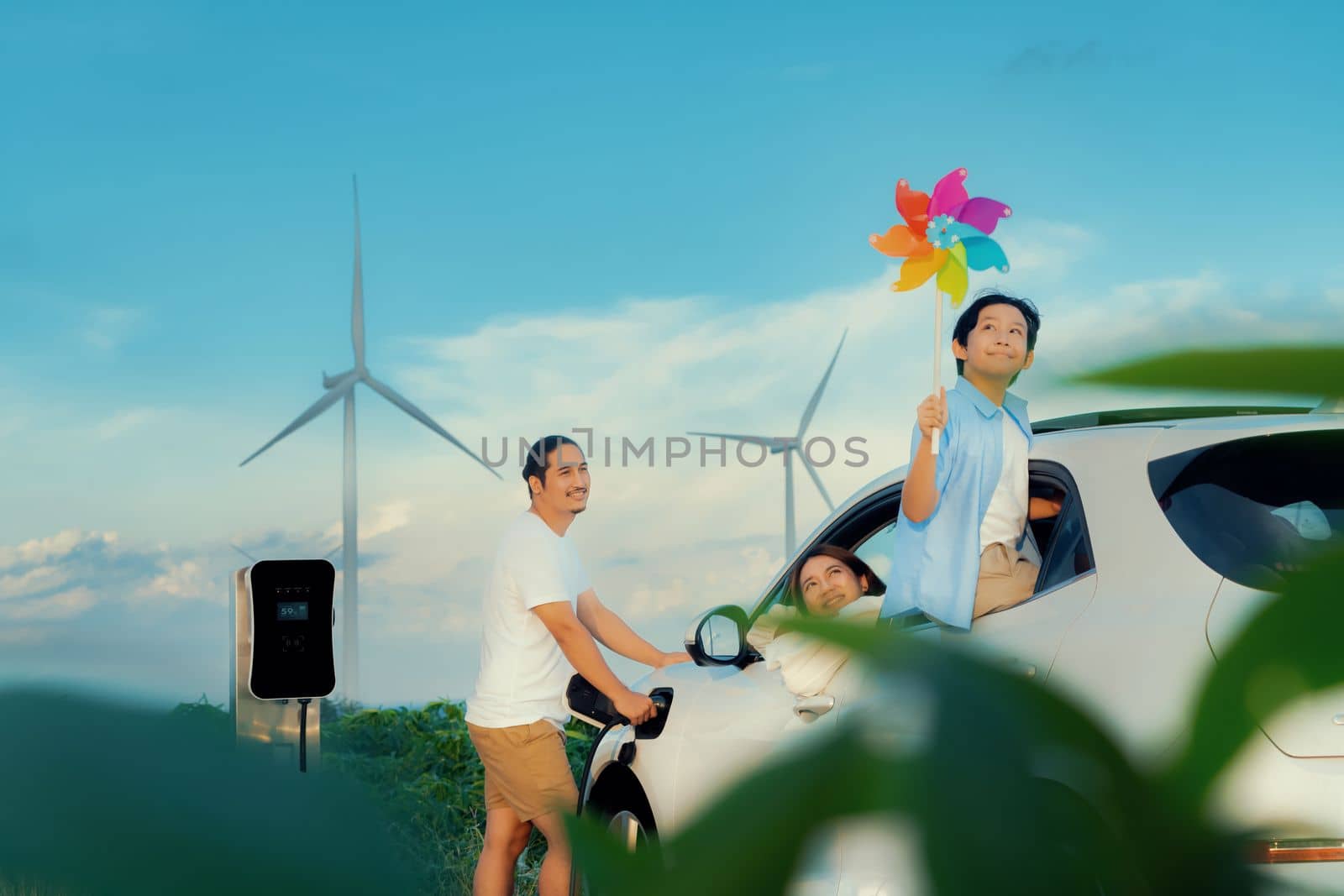 Concept of progressive happy family holding windmill toy and relax at wind farm with electric vehicle. Electric vehicle driven by clean renewable energy from wind turbine generator to charger station.