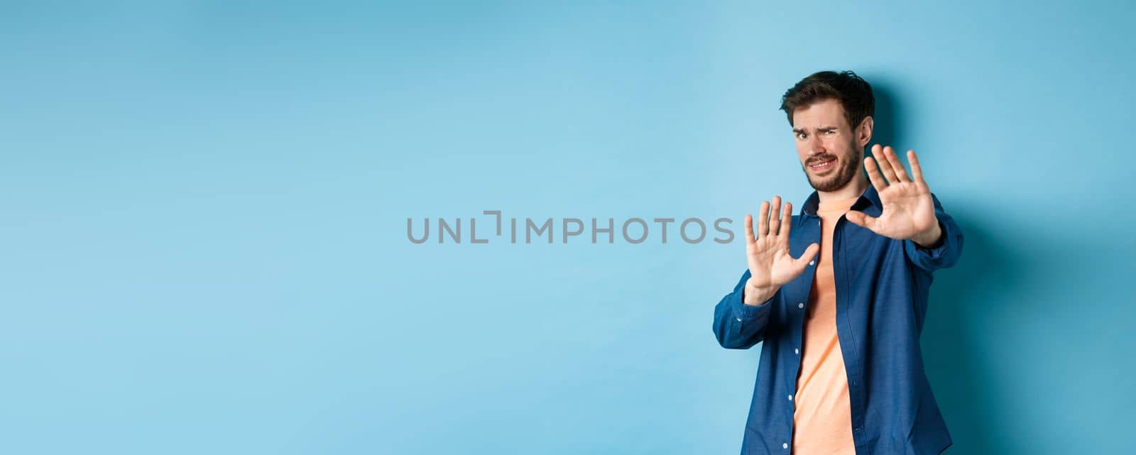 Guy cringe from something disgusting or embarrasssing, stretching out hands and asking to stop, grimacing displeased, standing on blue background by Benzoix