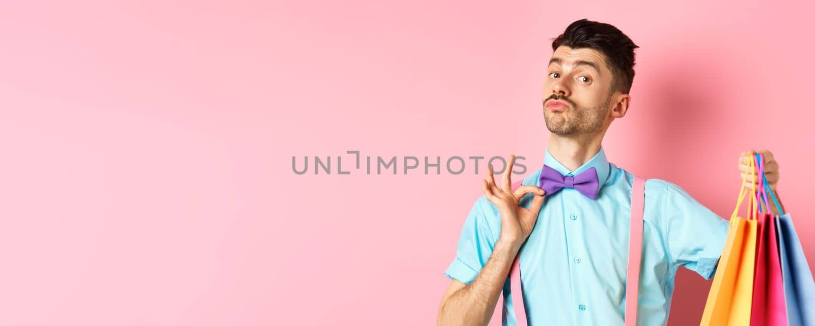 Fancy guy with moustache fixing his bow-tie and holding shopping bags, boyfriend bring gift packages, standing over pink background.