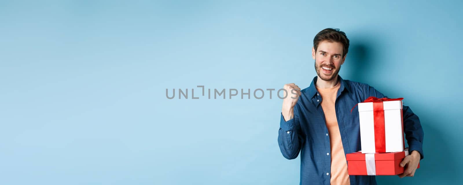 Cheerful young man got discounts on valentines day, making fist pump and say yes, holding gift boxes with presents for lover, standing over blue background by Benzoix