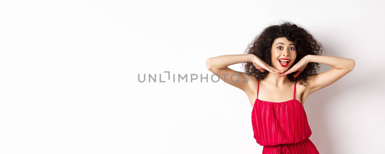 Beauty. Happy elegant woman with curly hair and makeup, wearing red dress, showing her face and smiling excited, standing against studio background by Benzoix