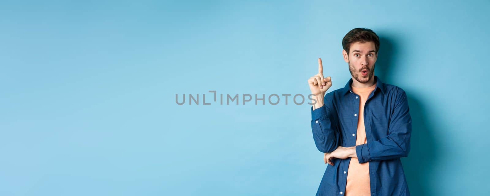 Handsome young man having an idea, pitching plan, raising finger and saying suggestion, standing on blue background by Benzoix