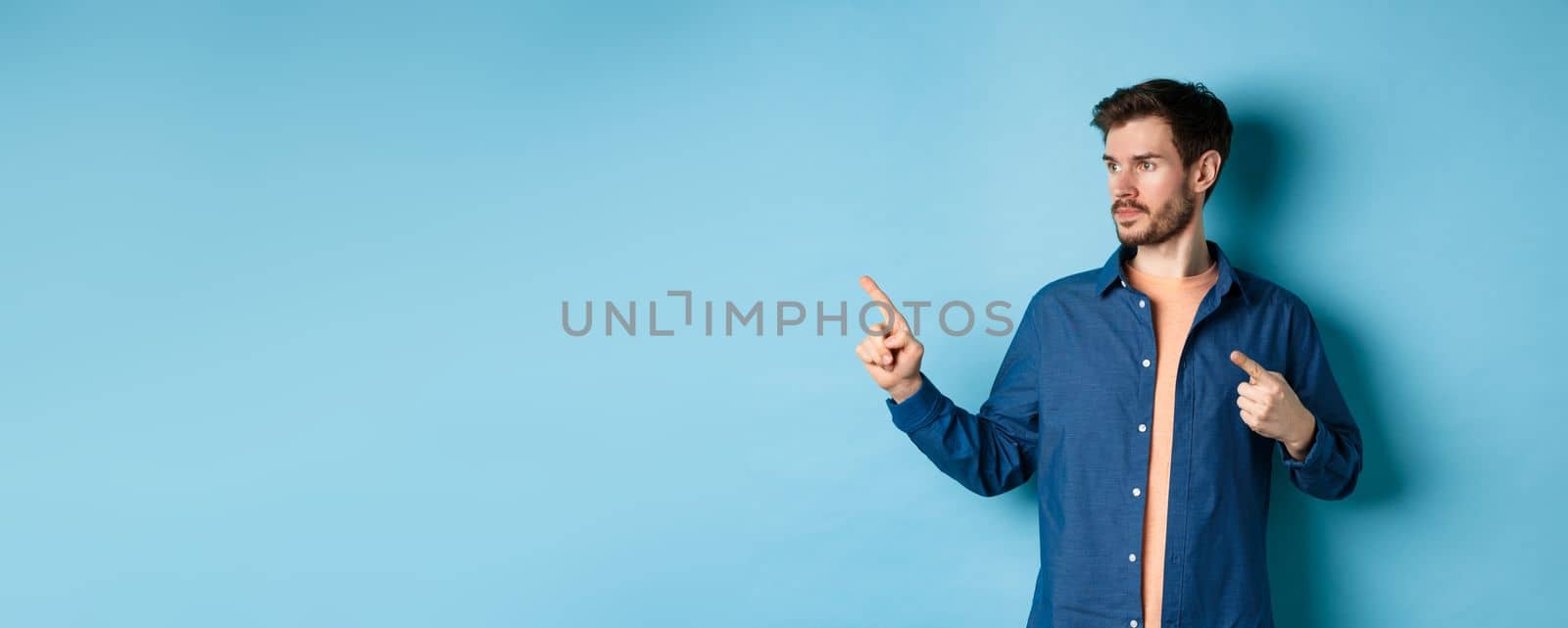 Image of young caucasian man looking and pointing aside, showing advertisement, standing on blue background by Benzoix