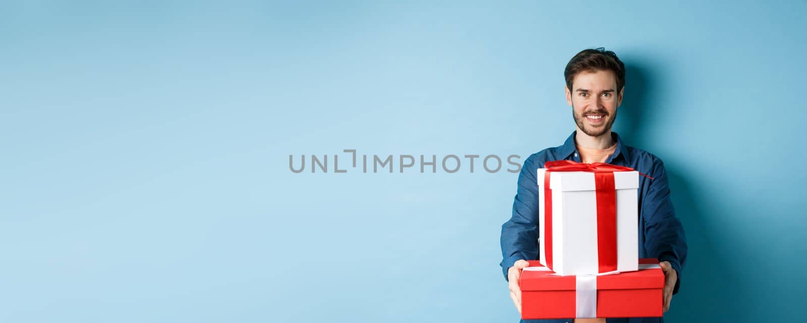Happy valentines day. Handsome man giving girlfriend presents, holding gift boxes and smiling, standing over blue background by Benzoix