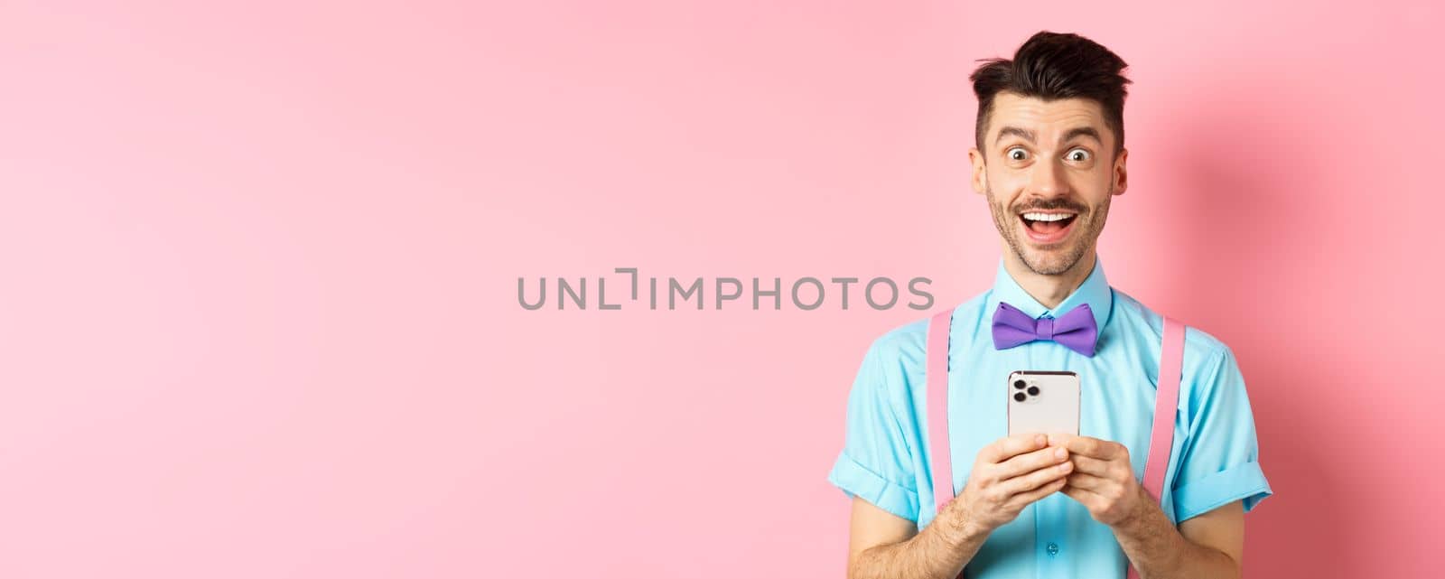 Online shopping. Happy man looking amazed after reading smartphone screen, smiling excited at camera, standing over pink background.