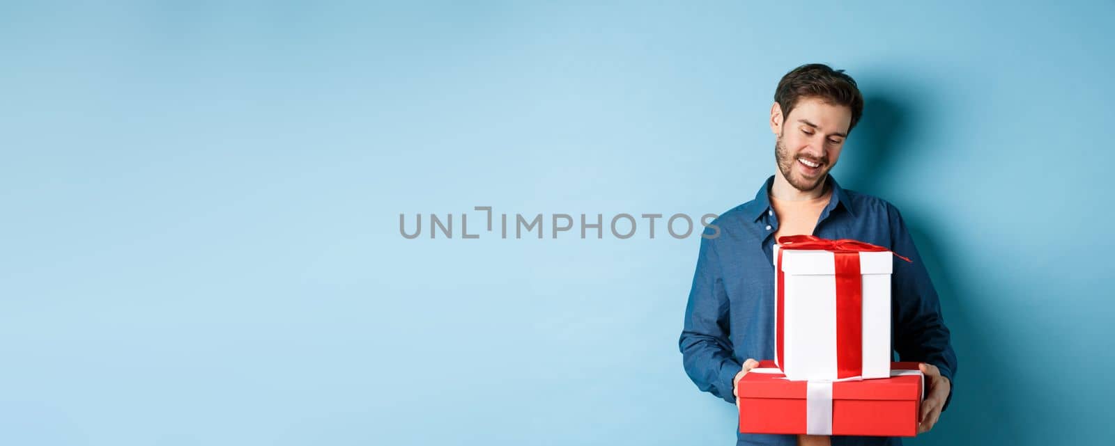Romantic young man with beard, looking happy at gift boxes on valentines day, giving presents to lover, standing over blue background by Benzoix