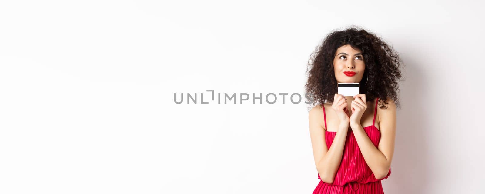 Silly young woman looking romantic, holding plastic credit card and thinking of shopping, standing over white background. Copy space
