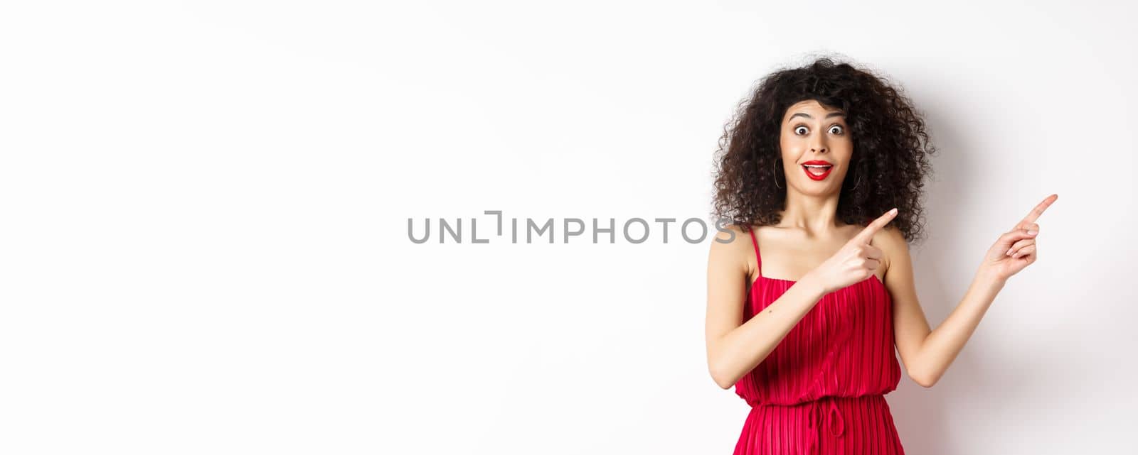 Surprised woman with curly hair, makeup and red dress, looking amazed and happy while pointing fingers right at logo, showing advertisement, white background.