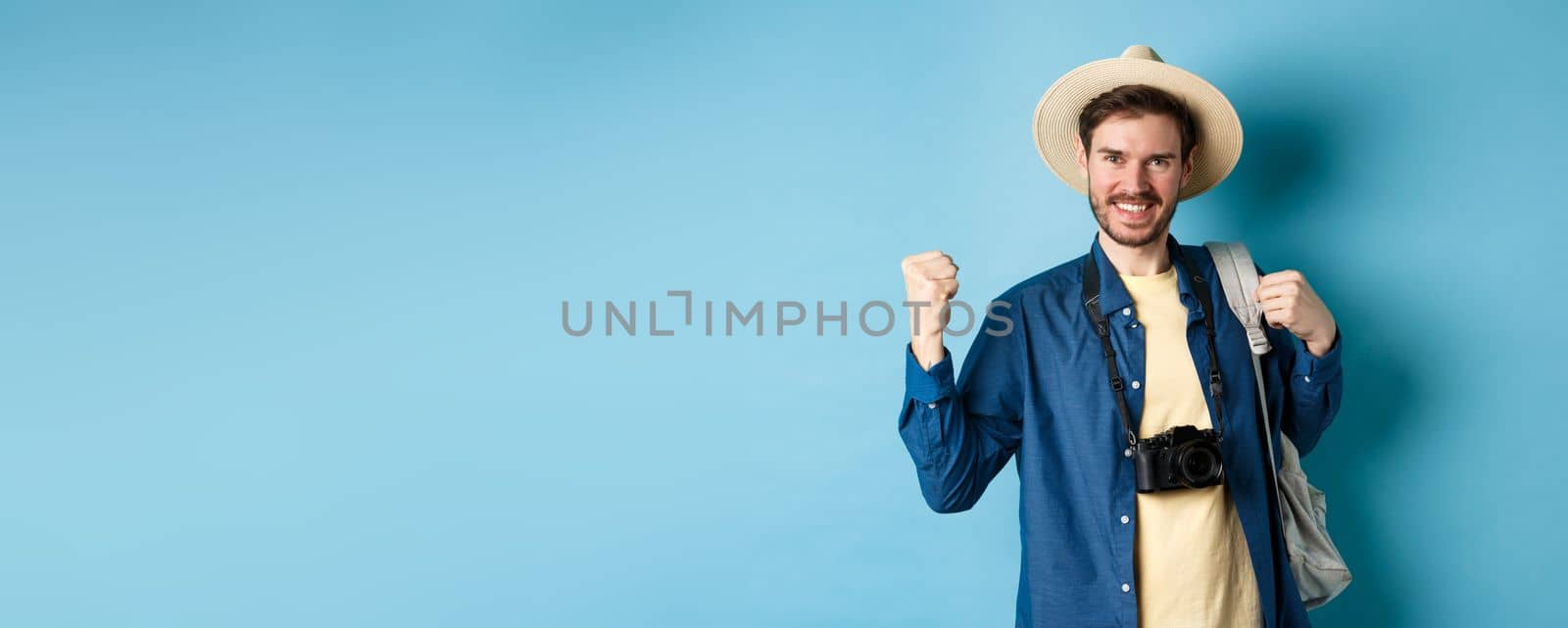 Happy tourist enjoying summer vacation, smiling and saying yes with fist pump, celebrating on holidays, feeling joy as finaly travel abroad, standing on blue background by Benzoix