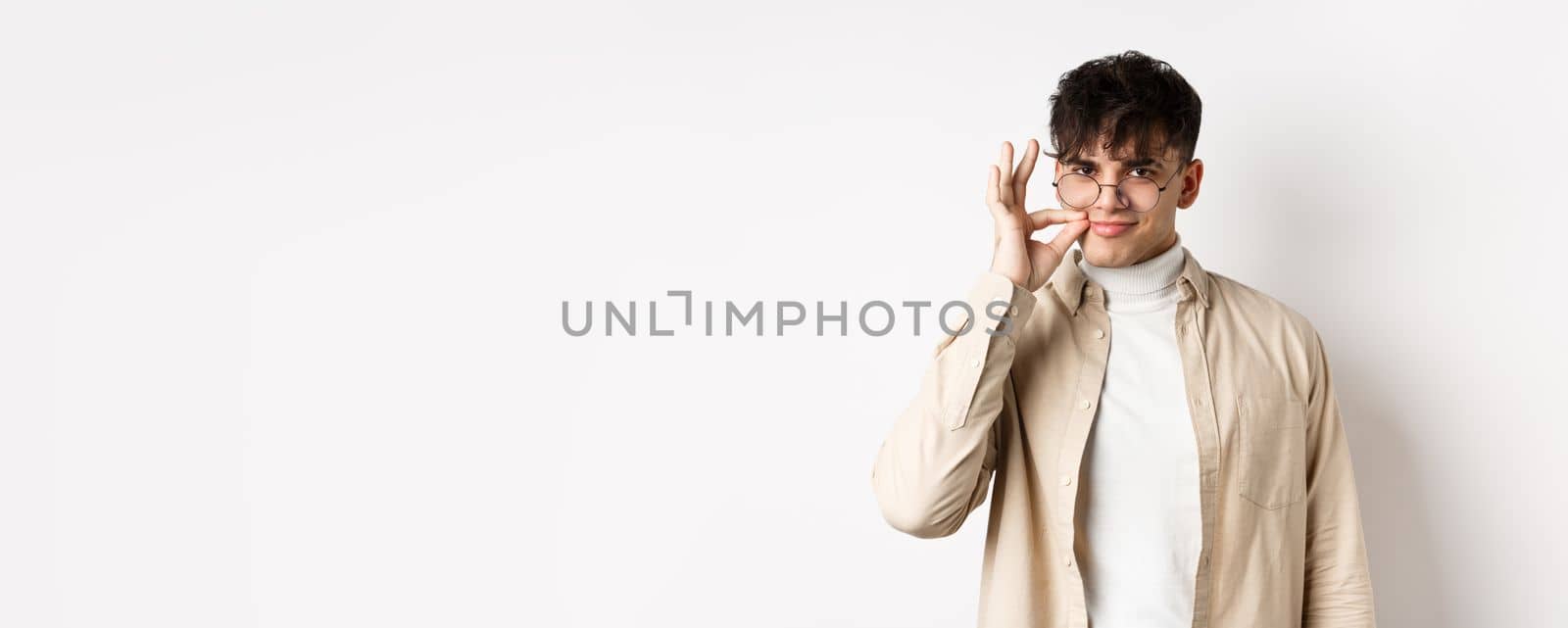 Smiling cunning guy in glasses shut his mouth on zipper, showing seal gesture near lips and smiling devious, keeping a secret, standing on white background.