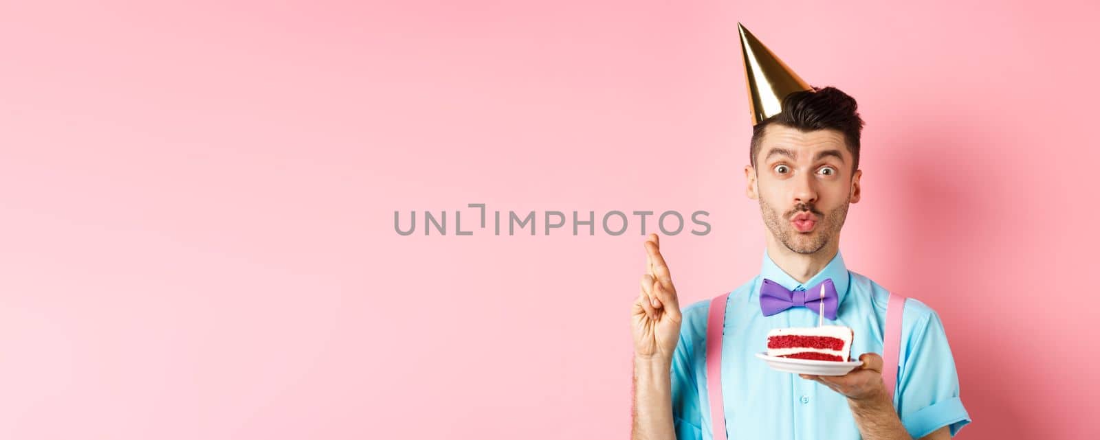 Holidays and celebration concept. Cute and funny man making birthday wish with crossed fingers, holding b-day cake and looking dreamy at camera, pink background.