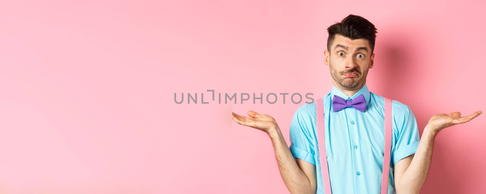 Image of confused guy in bow-tie and suspenders know nothing, shrugging shoulders and looking clueless, standing over pink background.