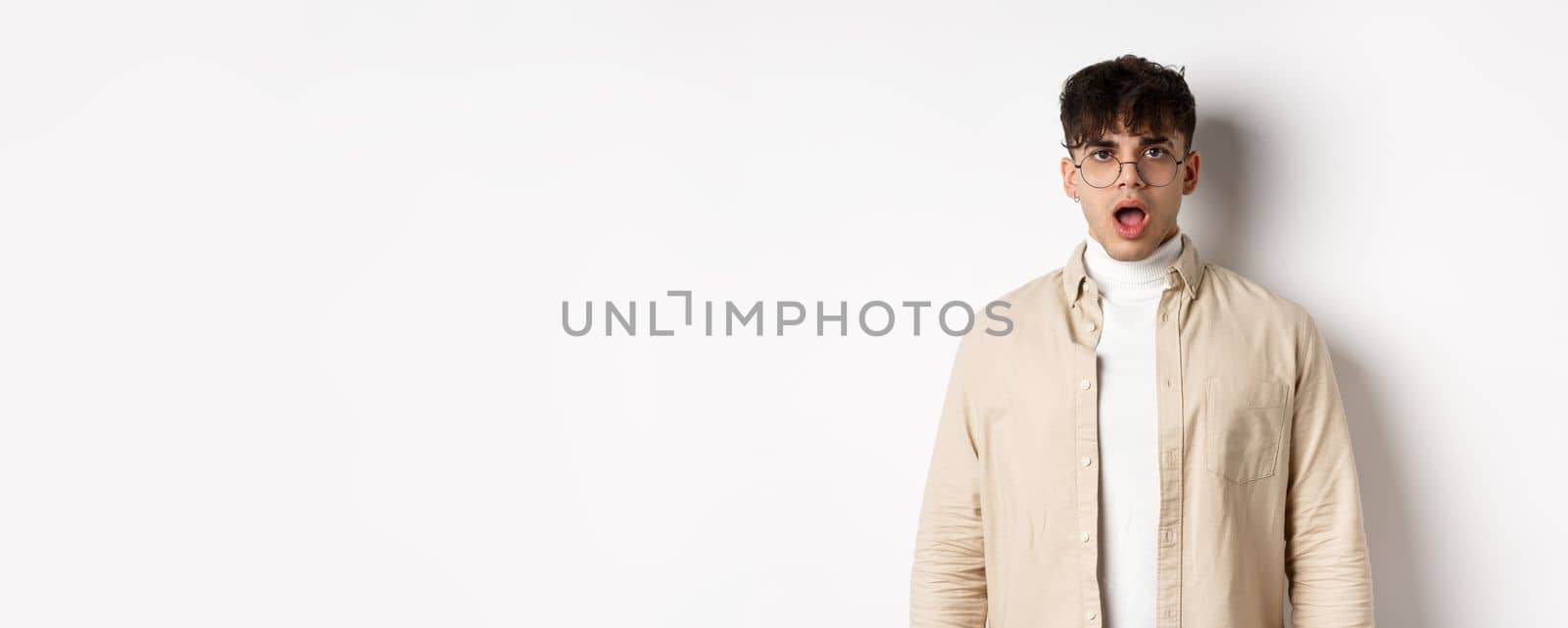 Portrait of shocked young man in glasses drop jaw, open mouth and stare amazed at camera, looking at something impressive, standing on white background.