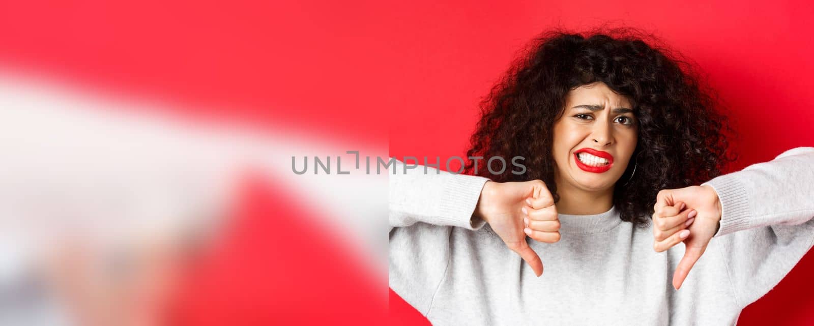 Close-up portrait of disappointed woman grimacing, cringe from something bad, showing thumbs-down, dislike and disapprove, standing on red background.