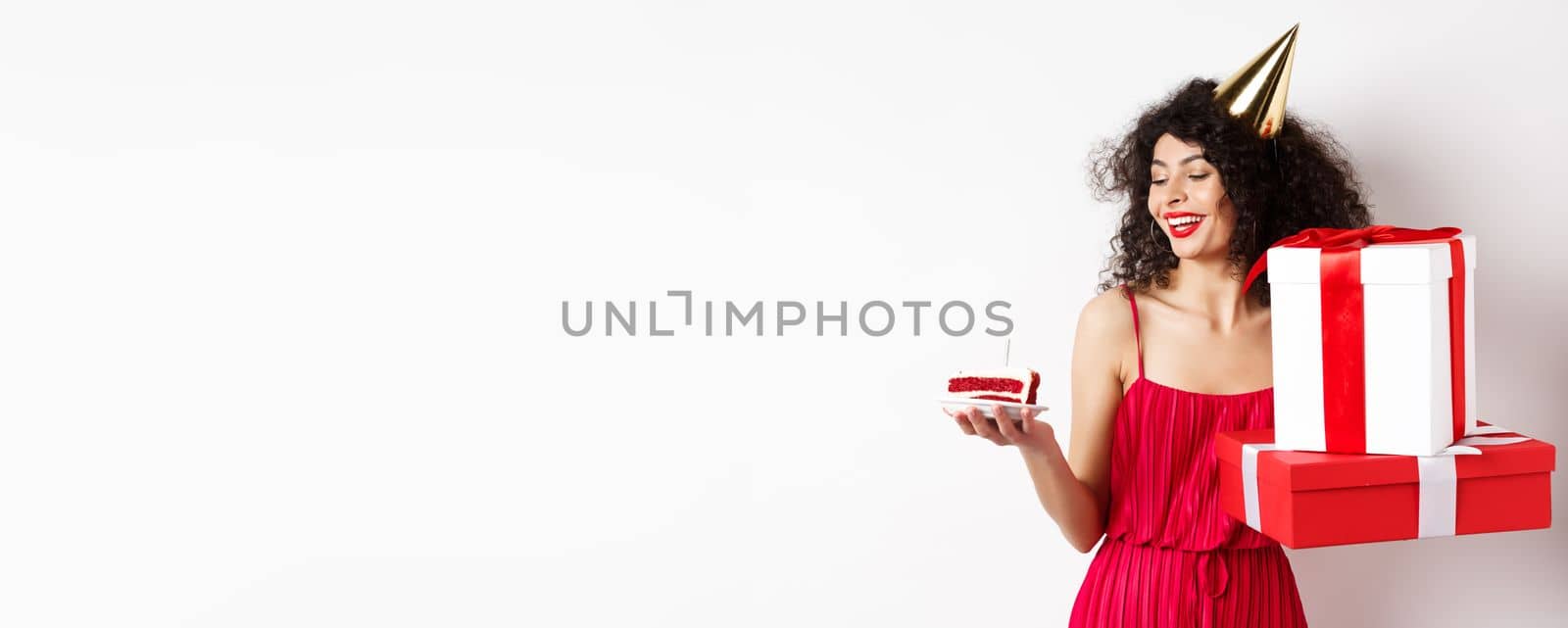 Cheerful smiling woman in red dress and party cone, looking happy at birthday cake, holding gifts, standing on white background. Copy space