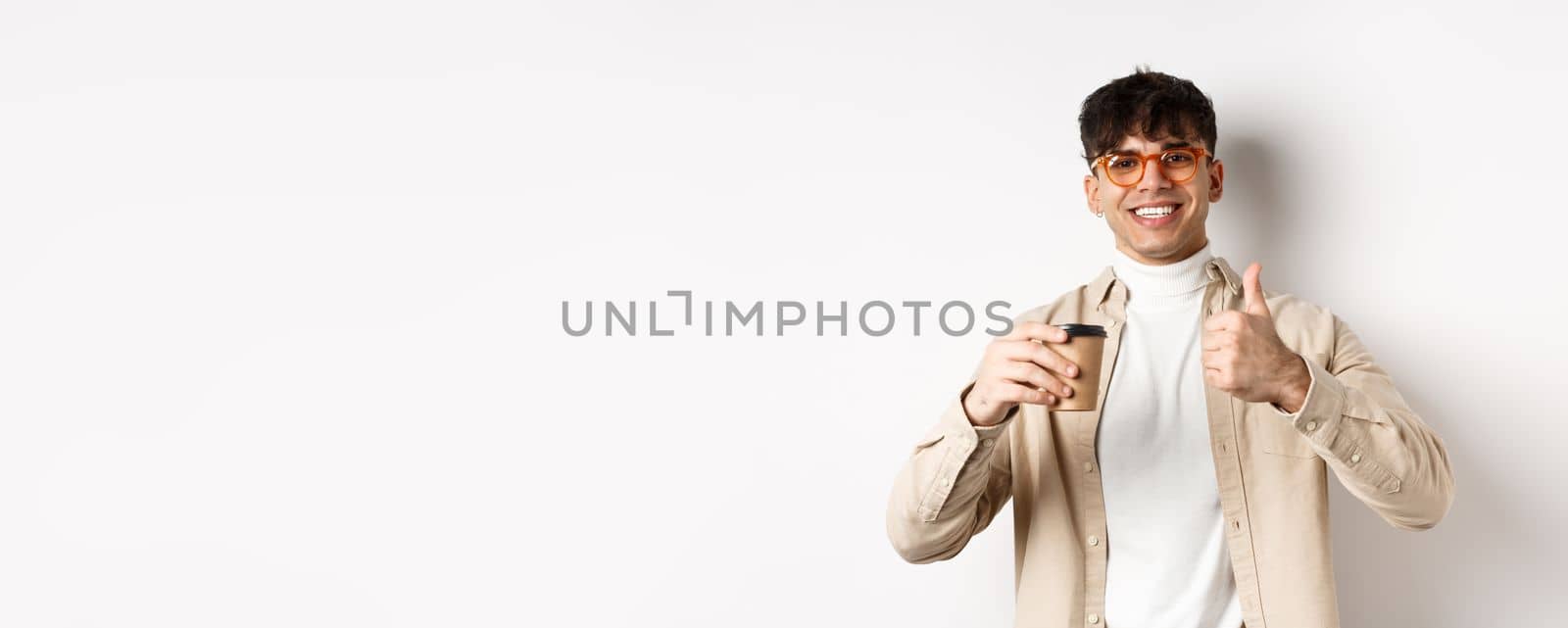 Real people. Cheerful smiling guy in glasses drinking coffee and showing thumb up, recommending cafe, standing on white background.