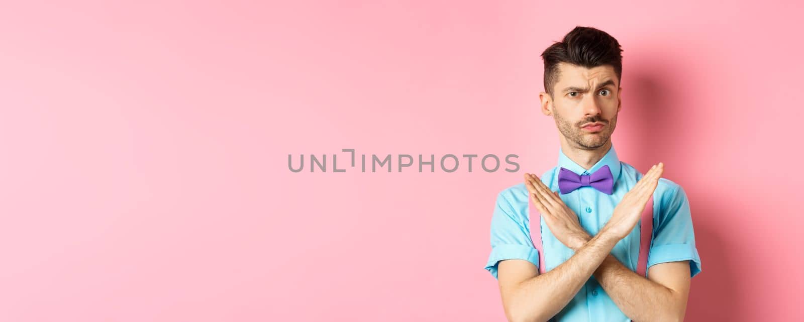 Serious-looking skeptical guy showing cross sign to stop, prohibit something bad, forbid action, standing on pink background. Copy space