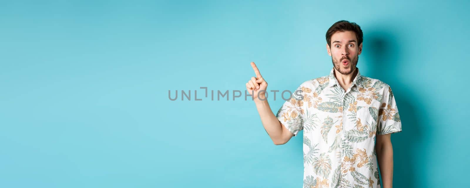 Surprised and amazed tourist in hawaiian shirt pointing finger left, showing promo and staring at camera, standing on blue background.