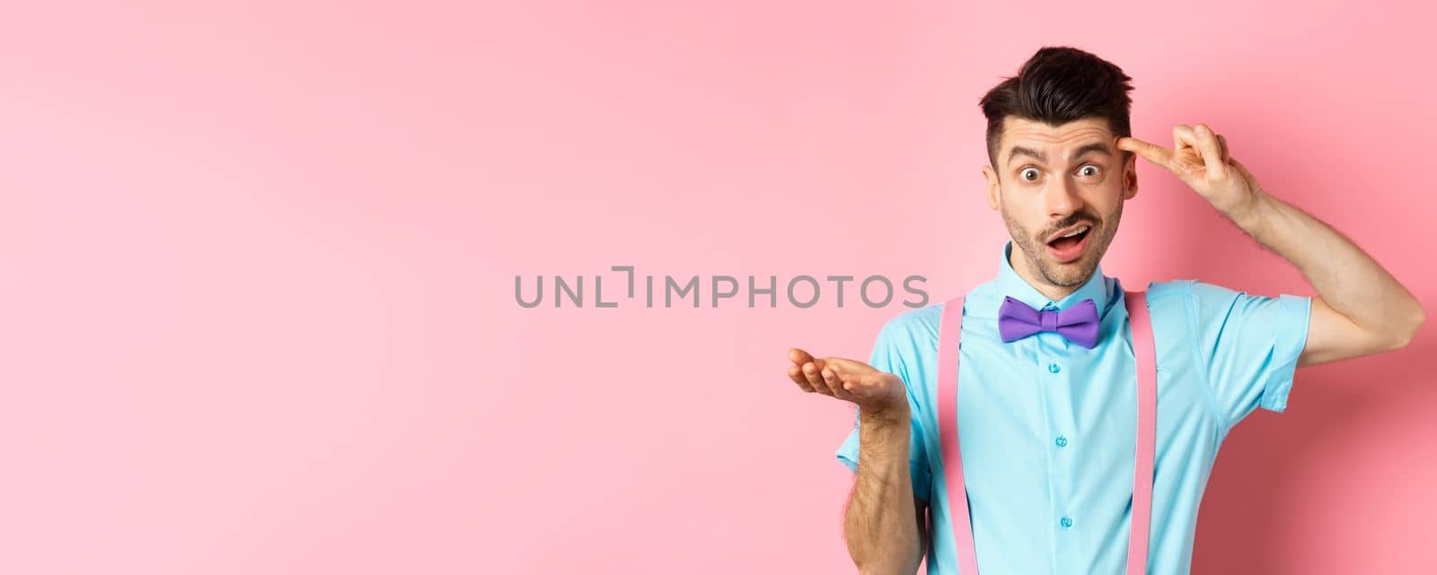 Annoyed guy mocking someone stupid, pointing finger at head and camera, are you stupid gesture, standing over pink background.