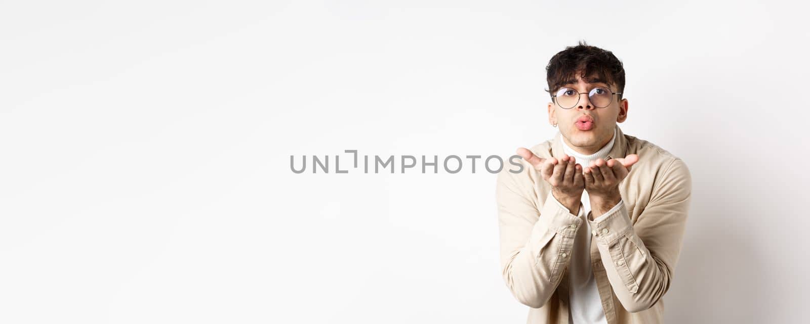 Cute boyfriend sending air kiss at camera on happy Valentines day, looking tender at lover, standing in casual clothes and glasses on white background by Benzoix