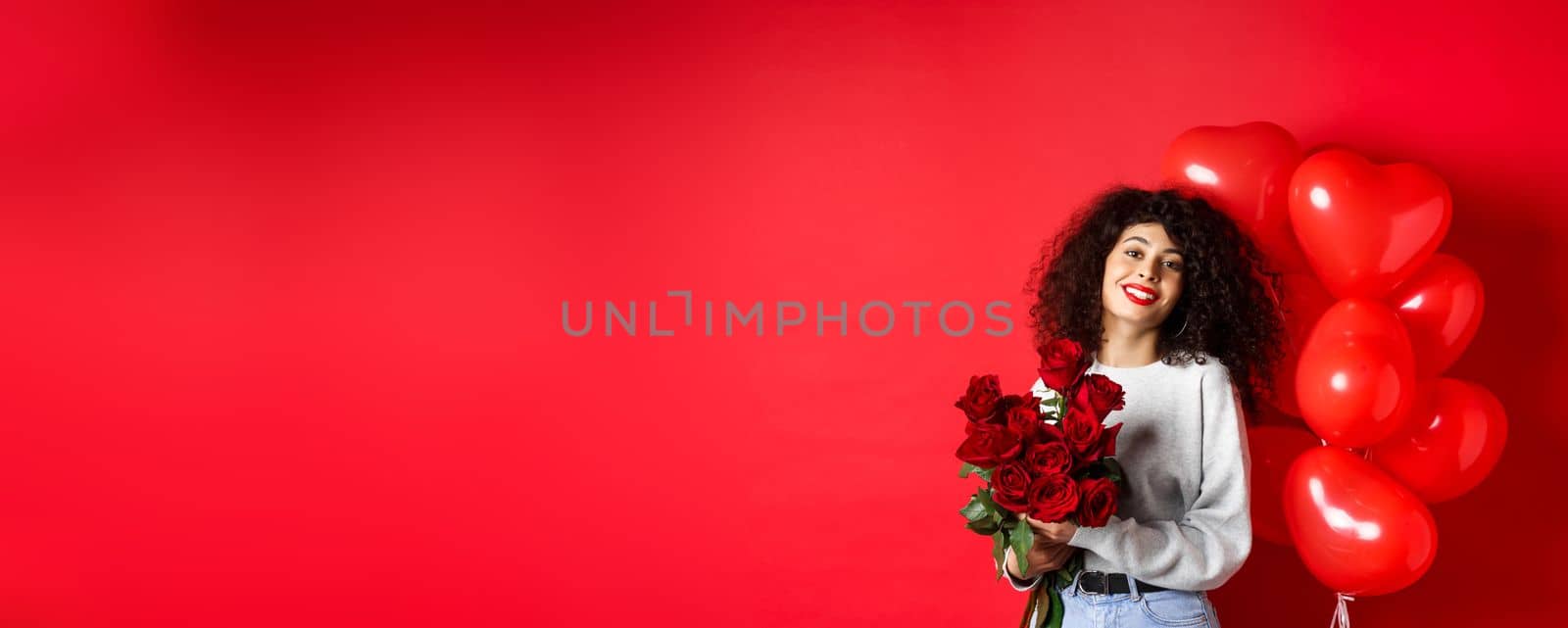 Holidays and celebration. Happy beautiful woman with curly hair, receive bouquet of roses and smiling, standing near party balloons, red background.
